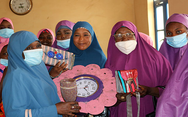 A group of women in pink and purple robes, protected by corona masks are holding two self-made colorful bags, a clock, and a wine cup made of recycled plastic trash in a room in Nigeria. 