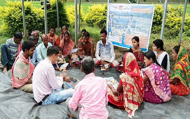 A sensitisation Program consisting of both men and women farmers organized for training on operation of solar pumps and subsidy schemes in East India. The group sits on the floor in a circle on a colourful blanket, wearing traditional Indian clothing and listening to a trainer sitting in the middle of the group.