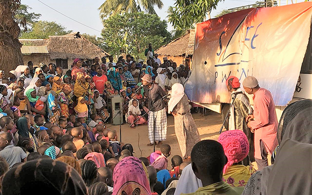 A large group of a village community in Kenya is gathered to listen to two people talking in the middle of the group into microphones. In the background is a large banner of S.A.F.E- the projects implementing organisation.