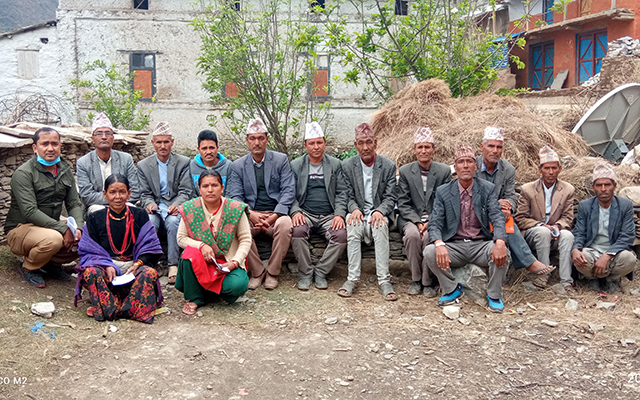 In the picture we see a group of 12 men and two women sitting crouched and looking towards the camera. The men are wearing traditional Nepalese caps. 