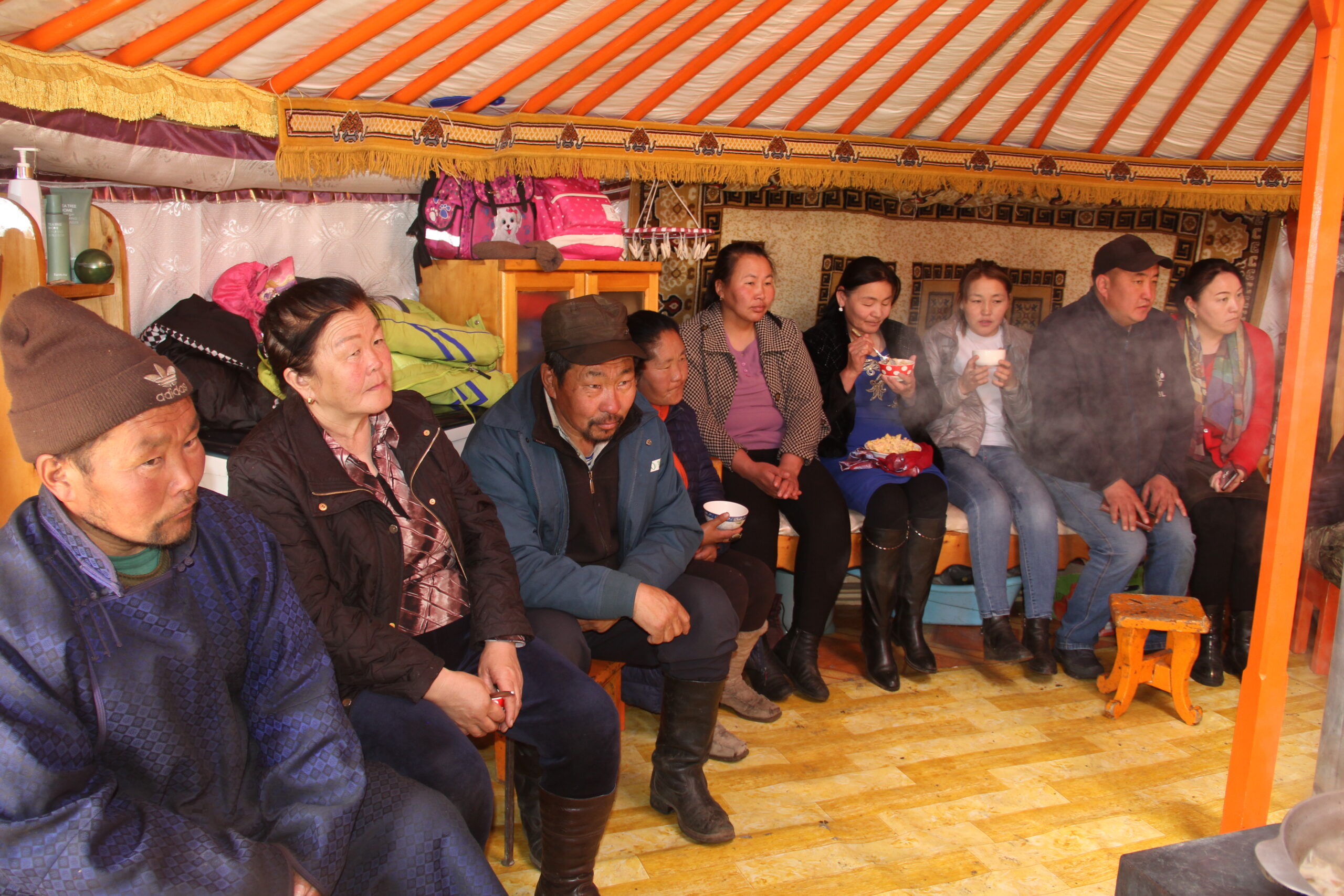 Several women and men sitting in a wooden round tent. They are holding tea cups and listening t a person that is not in the picture.
